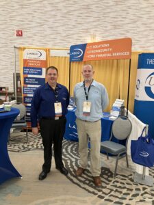 Two men stand in a convention center surrounded by marketing materials.