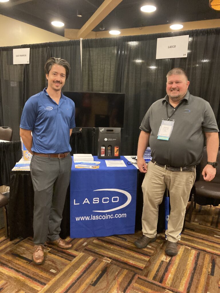 Two men stand in front of marketing materials in a convention setting.