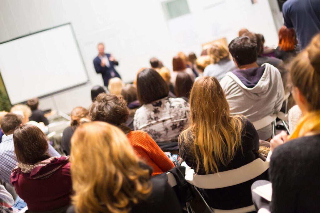 Person speaking at the front of a room with seated audience.